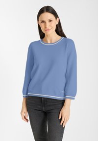 Vrouw met lang donker haar, gekleed in een lichtblauwe trui met witte accenten en zwarte jeans, staand tegen een plain witte achtergrond.