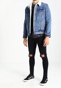 Veste en denim avec col en fourrure, couleur bleue, poches avant ; associée à un jean noir déchiré et des baskets noires avec des accents blancs.