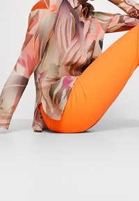 Blouse semi-transparente à motif floral dans des couleurs pastel associée à des leggings ajustés orange vif ; texture lisse, manches longues, design à boutons.