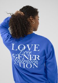 Femme aux cheveux bouclés attachés, portant un sweat-shirt bleu avec du texte blanc superposé "Love Generation" dans le dos, de dos.