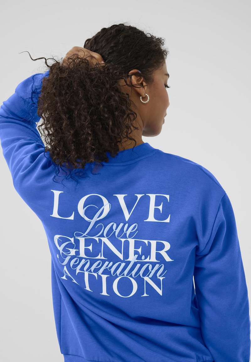 Femme aux cheveux bouclés attachés, portant un sweat-shirt bleu avec du texte blanc superposé "Love Generation" dans le dos, de dos.