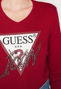 Röd långärmad T-shirt med V-ringning, som har en triangulär vit "GUESS"-logotyp och en svart glittrande "ICON"-design i strass.