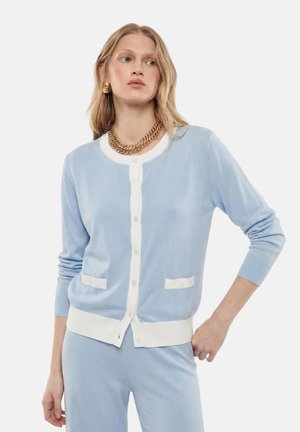 Donna che indossa un cardigan azzurro chiaro con bordo e bottoni bianchi, pantaloni coordinati, collana a catena dorata e orecchini dorati, posando con la mano sul fianco.