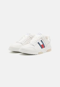 Sneakers bianchi con punta perforata, dettagli nelle strisce blu e rosse, pannelli laterali texturizzati e suola in gomma beige. La linguetta presenta un dettaglio con logo.
