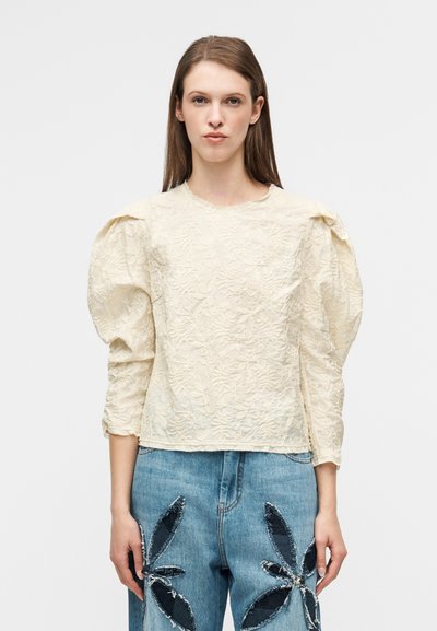 Κρεμ μπλούζα με υφή, φουσκωτά μανίκια και λεπτομέρειες με λουλουδάτο σχέδιο, συνδυασμένη με τζιν παντελόνι που έχει σκοτεινά λουλουδάτα εφαρμογές.