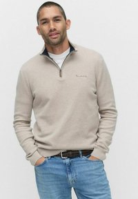 Beige zip-up tröja med ståkrage, strukturerad tyg och en liten logotyp på bröstet, bärs över en vit T-shirt och blå jeans.