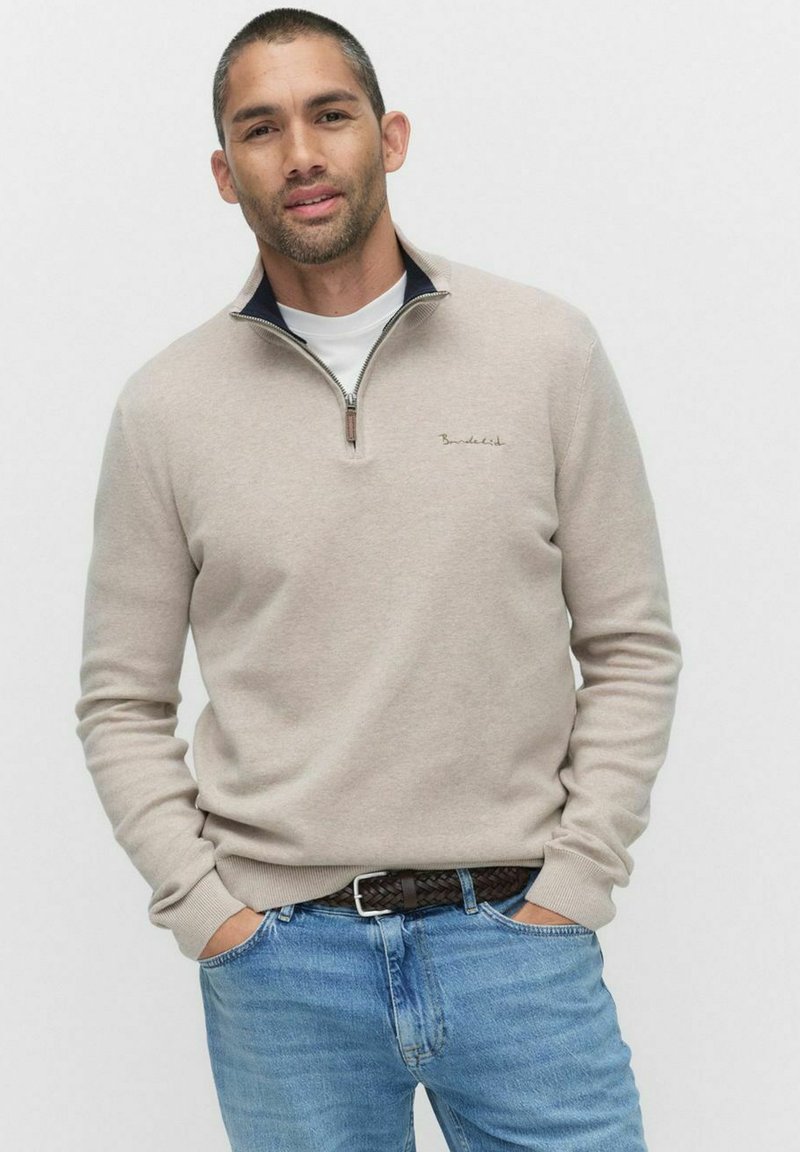 Beige zip-up tröja med ståkrage, strukturerad tyg och en liten logotyp på bröstet, bärs över en vit T-shirt och blå jeans.