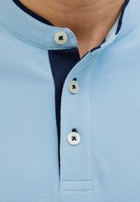 Polo in azzurro chiaro, in tessuto testurizzato. Presenta un colletto blu navy con due bottoni. Gli accenti dei bottoni sono bianchi con un design a contrasto.