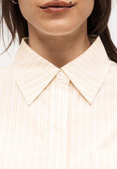 Γυναίκα με κρεμ πουκάμισο με λεπτές ρίγες και γιακά, κουμπωμένο στο πάνω μέρος, με ορατό το κάτω μέρος του προσώπου και ελιά πάνω από το χείλος.