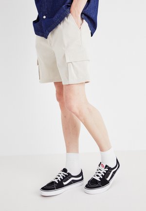 Shorts cargo en velours côtelé beige avec poches latérales, associés à des baskets noires avec des accents blancs et des lacets, portés avec des chaussettes blanches montantes.