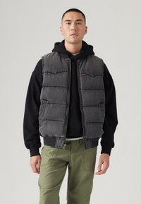 Sivi puff jacket s patentnim zatvaračem, bez rukava, s kvadratnim dijelovima, složen preko crne dukserice, u kombinaciji s maslinasto zelenim hlačama.