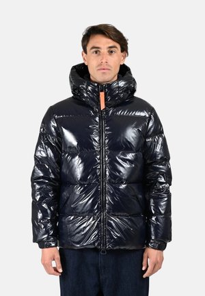 Homme portant une doudoune noir brillante avec capuche, fermeture éclair à l'avant et mains détendues sur les côtés, sur un fond léger uni.