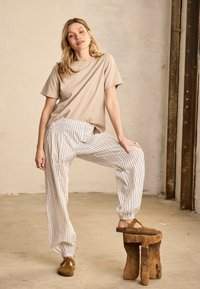 Fiatal nő bézs pólóban, fehér csíkos nadrágban és barna bebújós cipőben, egyik lába egy rusztikus fa székpárkányon pihen.