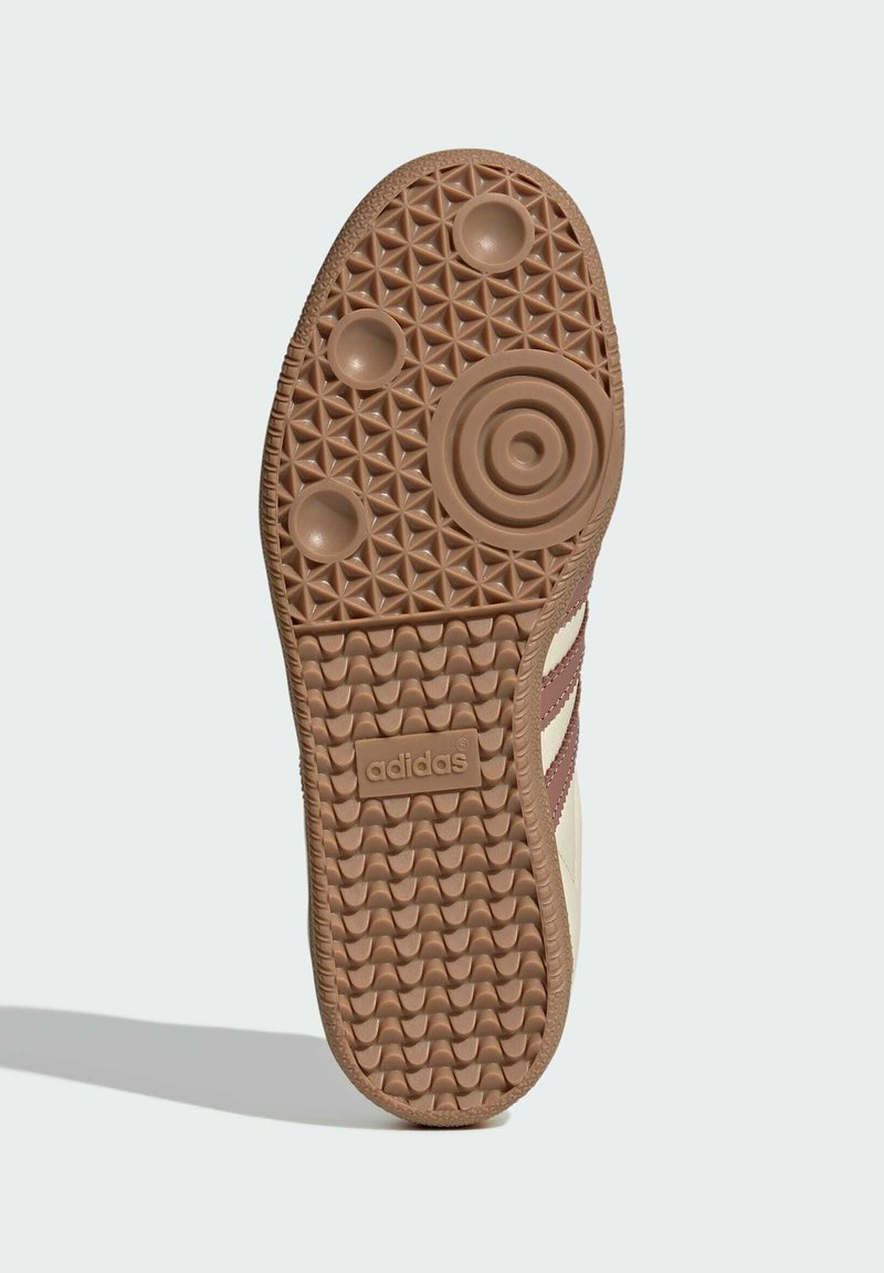 Brun gummiyttersida med texturerat mönster, cirkulära indragningar och upphöjd logotyp. Beige överdel med mörka ränder och kontrasterande detaljer.