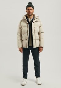 Lichtbeige pufferjack met capuchon, met een gladde textuur en drukknoopsluiting. Draag het over een donker shirt en marineblauwe broek, met witte sneakers.
