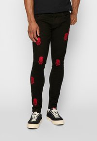 Tievās melnās bikses ar sarkano kaulu un galvaskausa izšuvumu visā virsmā. Iespējas ietver tradicionālo piecu kabatu dizainu un vieglu mirdzumu.