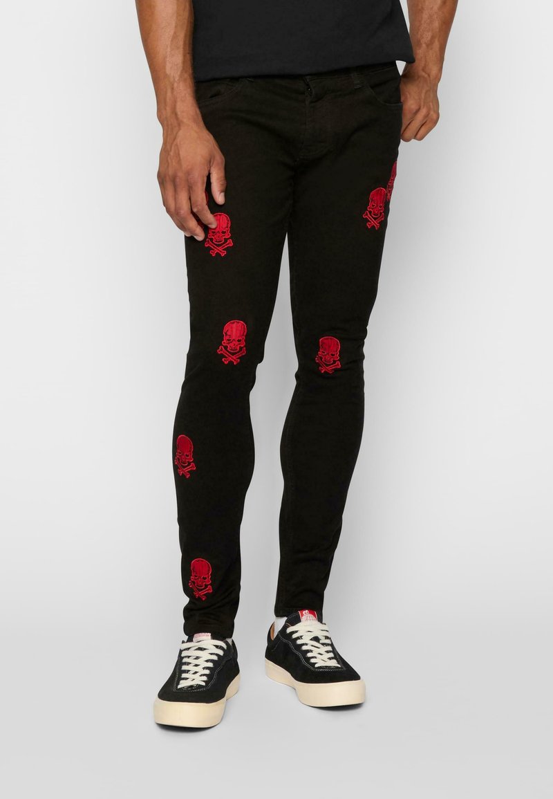 Tievās melnās bikses ar sarkano kaulu un galvaskausa izšuvumu visā virsmā. Iespējas ietver tradicionālo piecu kabatu dizainu un vieglu mirdzumu.