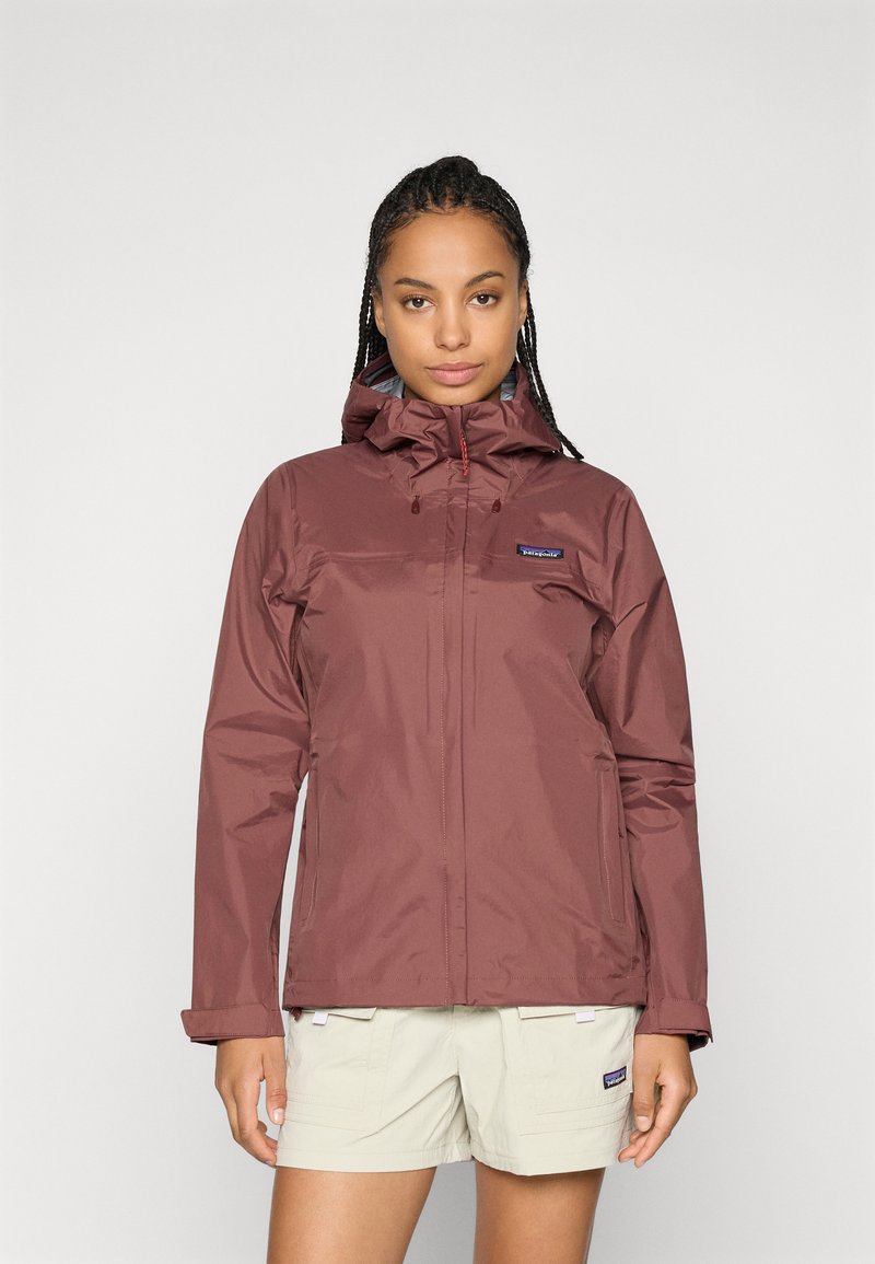 Vrouw met gevlochten haar, gekleed in een kastanjebruine hoodie en beige shorts, staand tegen een effen lichte achtergrond.