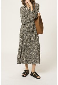 Donna che indossa un abito midi a maniche lunghe nero e beige con un motivo, sandali neri intrecciati, mentre porta una grande borsa a spalla in pelle marrone.