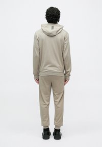 Beige hettegenser og matchende joggebukser laget av mykt stoff. Har hette bak, ermer med mansjetter og bukser med smale ankler. Sorte sko.