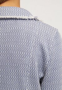 Blå och vit mönstrad tyg med en texturerad väv, med fransade kanter på kragen. Designen har ett geometriskt mönster.