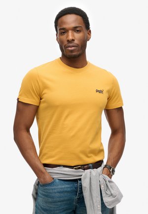 Man wearing a mustard yellow t-shirt, blue jeans, watch on left wrist, and light striped shirt tied around waist against white background.