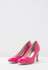 Skimrande ljusrosa högklackade skor med spetsiga tår och beige inredning på en vit yta med enkel bakgrund.