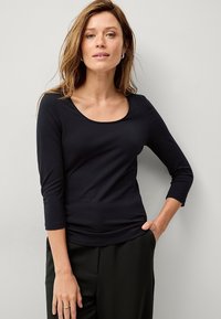 Schwarzes, langärmliges Oberteil mit runder Ausschnitt. Glattes, elastisches Material. Figurbetonter Schnitt, kombiniert mit dunklen Hosen. Einfach und vielseitig.