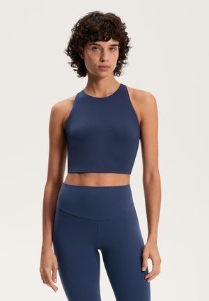 Frau mit kurzen lockigen Haaren, die ein marineblaues ärmelloses Crop-Top und passende hochgeschnittene Leggings trägt, steht vor einem einfarbigen Hintergrund.