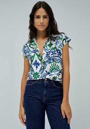 Blusa de manga corta en blanco con patrones florales en azul y verde, con cuello y botón en la parte delantera, combinada con jeans de un azul oscuro.