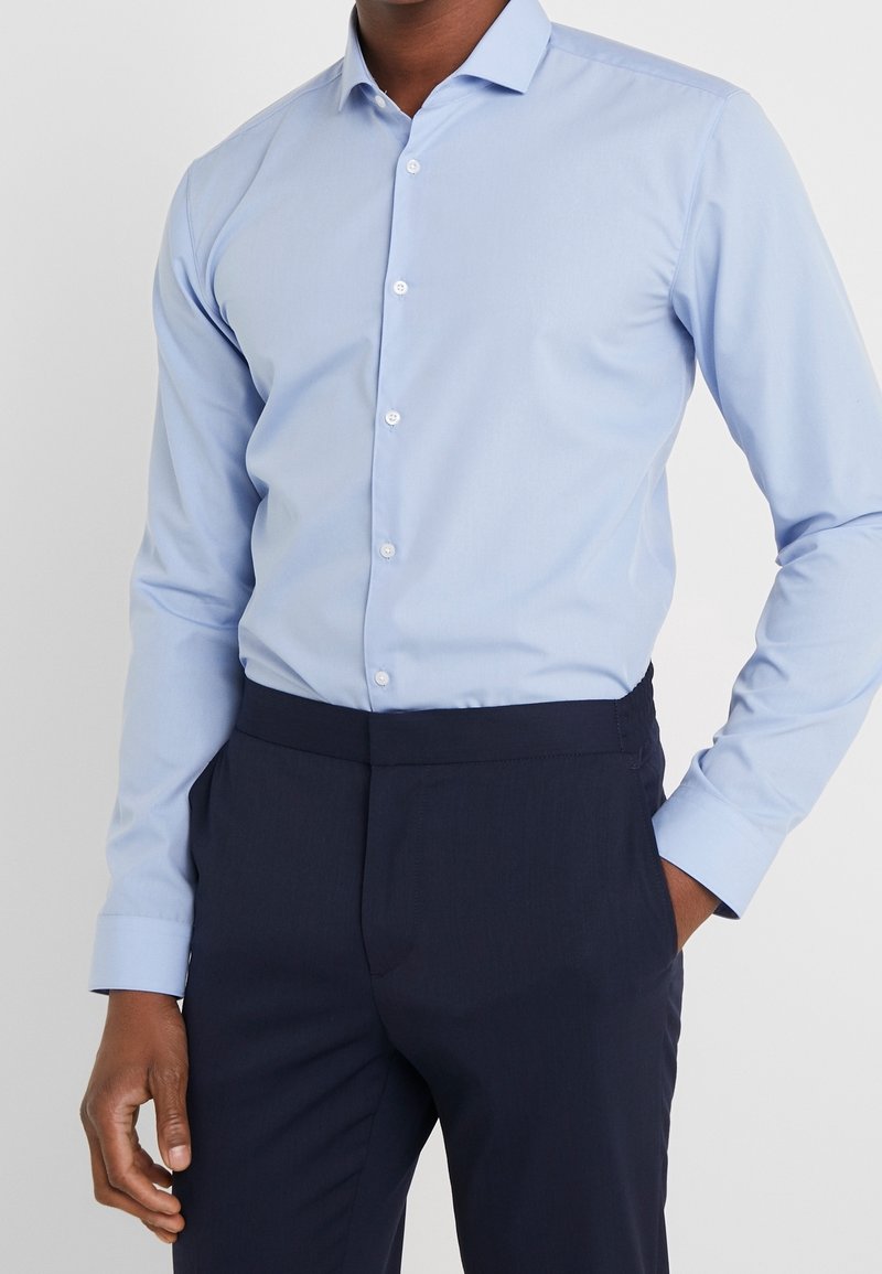 Homme portant une chemise bleu clair boutonnée rentrée dans un pantalon bleu marine foncé, une main dans la poche et fond neutre.