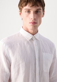 Camisa de linho rosa claro, com botões, colarinho clássico, um único bolso no peito e uma textura subtil. Apresenta botões brancos ao longo da frente.