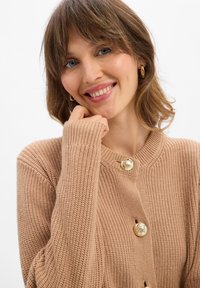 Frau mit hellbraunen Haaren, die goldene Creolen und eine beigen Strickjacke mit goldenen Knöpfen trägt, lächelt mit einer Hand nah am Kinn.
