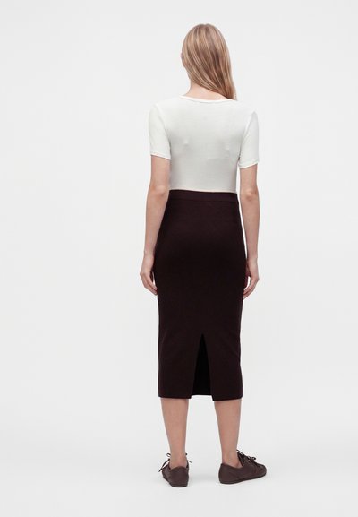 Femme debout, dos tourné, portant un haut blanc à manches courtes, une jupe midi noire avec une fente au dos, et des chaussures marron à lacets sur un fond uni.