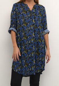 Blumige Tunika mit einem dunkelblauen Hintergrund, die blaue, gelbe und weiße Blumen zeigt, einen V-Ausschnitt hat und hochgekrempelte Ärmel besitzt. Gebogener Saum.