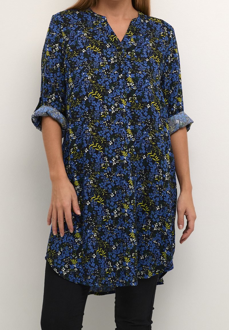 Blumige Tunika mit einem dunkelblauen Hintergrund, die blaue, gelbe und weiße Blumen zeigt, einen V-Ausschnitt hat und hochgekrempelte Ärmel besitzt. Gebogener Saum.