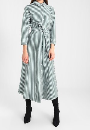 Vrouw draagt een midi-jurk met lange mouwen met groene en witte verticale strepen, een strik in de taille en zwarte enkellaarsjes met hak.