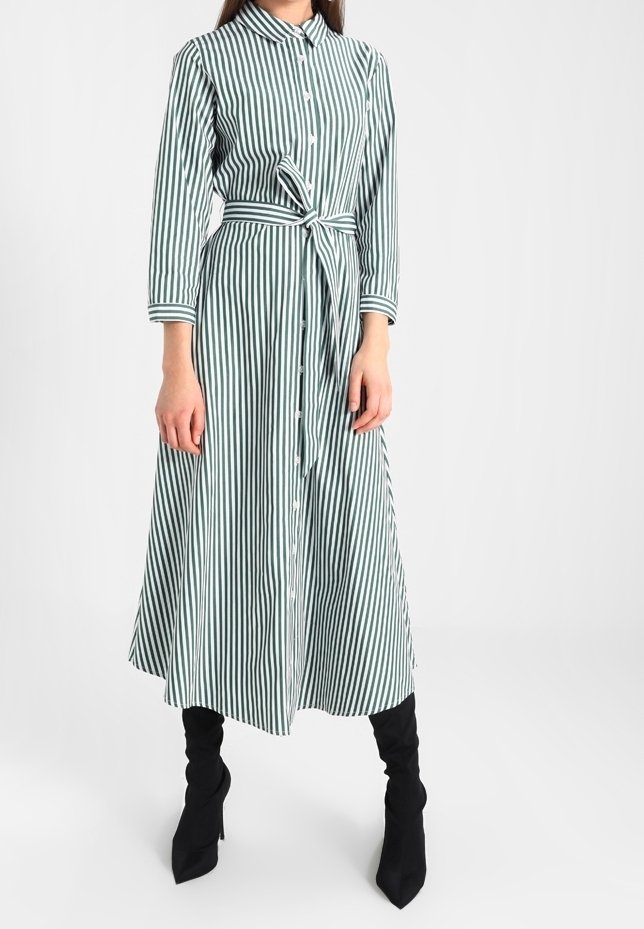Femme portant une robe midi à manches longues avec des rayures verticales vertes et blanches, une ceinture à la taille et des bottes noires à talons.