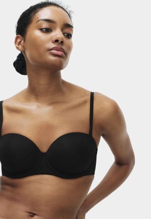 Frau mit geflochtenem Haar in einem schwarzen trägerlosen BH, die leicht nach unten und zur Seite blickt, vor einem schlichten hellen Hintergrund.