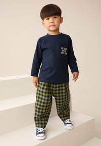 Temno modra majica z dolgimi rokavi z logotipom na prsih, kombinirana z zeleno-temno modrimi karirastimi ohlapnimi hlačami. Obutev so temno modri supergi.