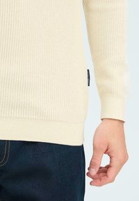 Lichtbeige geribde trui met lange mouwen, voorzien van een gestructureerde stof en een aansluitende manchette. Gecombineerd met donkerblauwe jeans.