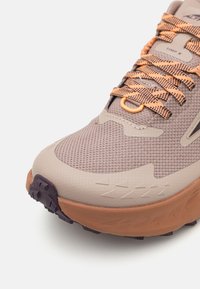 Chaussure de trail beige avec une tige en mesh, des lacets orange et noir, une semelle en caoutchouc et des accents texturés sur le côté et l'orteil.