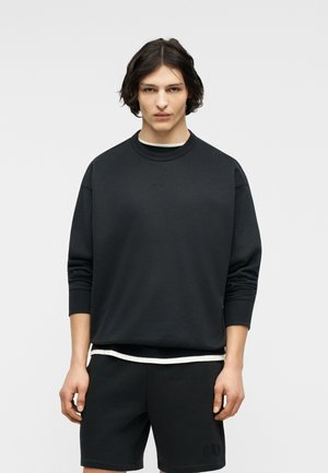 Mlada oseba s srednje dolgimi temnimi lasmi nosi črno jopa čez belo majico in črne kratke hlače, stoji ob enobarvni svetli ozadju.