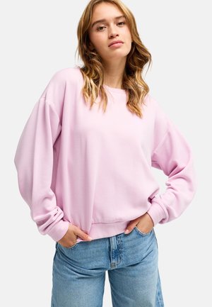 Jonge vrouw met golvend blond haar, draagt een losse lichtroze trui en blauwe spijkerbroek, staand met handen in de zakken tegen een witte achtergrond.