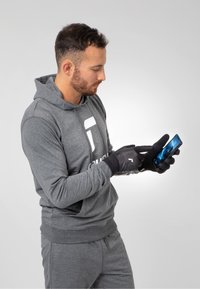 Grauer Hoodie mit einem Logo, graue Jogginghose und schwarze Touchscreen-Handschuhe. Die Person interagiert mit einem Smartphone, das ein Bild anzeigt.