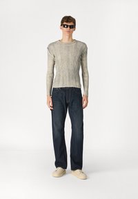 Svetlo siva rebrasta majica dugih rukava, temno modri jeans, in bež superge. Model nosi črne sončne očali in stoji pred enobarvno ozadjem.