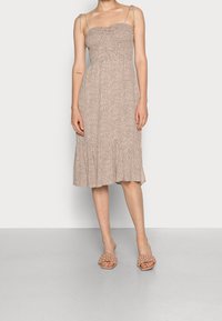 Robe midi à pois bruns avec corsage smocké et fines bretelles réglables. Jupe évasée avec ourlet volants. Associée à des sandales marron clair.