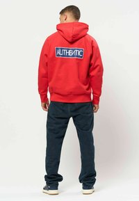 Homme portant un sweat à capuche rouge avec le texte "AUTHENTIC" dans le dos, jean foncé et baskets, debout de dos devant un fond uni.