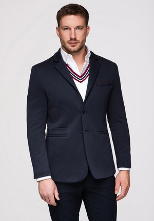 Mand iført navy blazer over hvid skjorte og hvid V-hals sweater med røde og navy striber, stående mod ensfarvet baggrund.