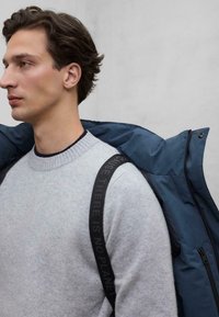 Jasnoszary sweter z prążkowanym dekoltem, noszony pod niebieską kurtką puchową. Czarny pasek z tekstem, widoczny na ramieniu. Gładka tekstura.